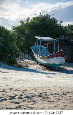 The picture shows a small fishing boat on the beach in front of a wooden hut and green bushes on a sunny day.