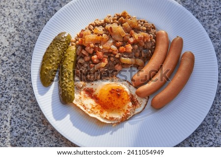 Close up picture on white porcelain plate with sweet and sour lentils mash, pork sausages, sunny side up egg and pickled cucumbers. Delicious, healthy dish with balanced nutrients and dietary fibre.