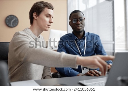 Waist up shot of young Caucasian male programmer pointing at laptop screen and talking to African American coworker while working in office