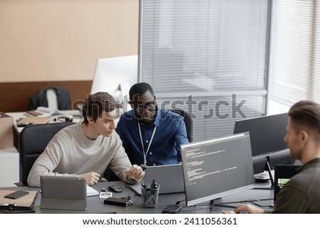 Wide angle shot of African American IT male mentor pointing out inaccuracies in code of young Caucasian male programmer while sitting with laptop at desk Royalty-Free Stock Photo #2411056361