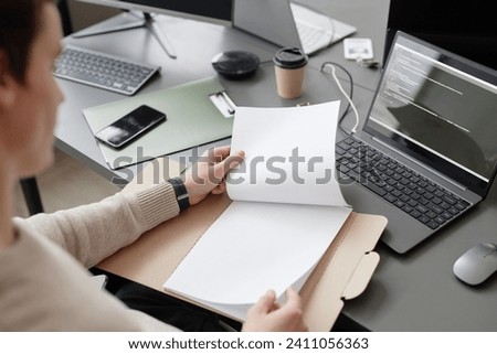Over the shoulder shot of unrecognizable IT worker holding blank papers in cardboard folder while sitting at table with computers