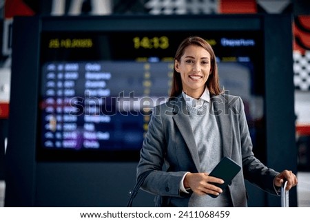 Young happy businesswoman standing at train station and looking at camera. Arrival departure board is in the background.