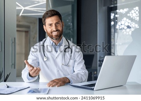 Portrait of a smiling young male doctor talking and consulting on a video call. He is sitting at the table in the office of the clinic, gesturing with his hand in front of the camera. Royalty-Free Stock Photo #2411069915