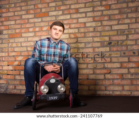 Young Handsome Man Sitting on Vintage Toy Automobile on Brick Wall Background While Looking at the Camera.