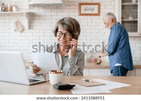 Busy european businesswoman old senior grandmother wife working, doing paperwork, dealing with documents while husband cooking dinner at home kitchen. Equality Royalty-Free Stock Photo #2411088951