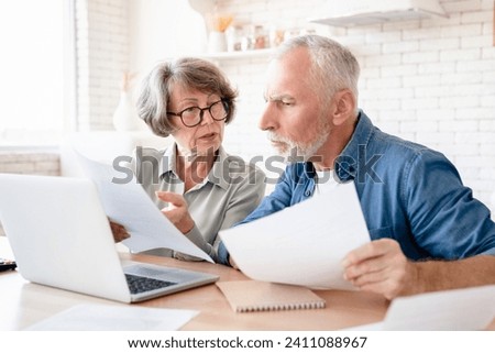 Concentrated concerned european senior old elderly couple spouses grandparents doing paperwork, paying domestic bills, rentals, receiving pension, calculating funds and budget at home kitchen Royalty-Free Stock Photo #2411088967