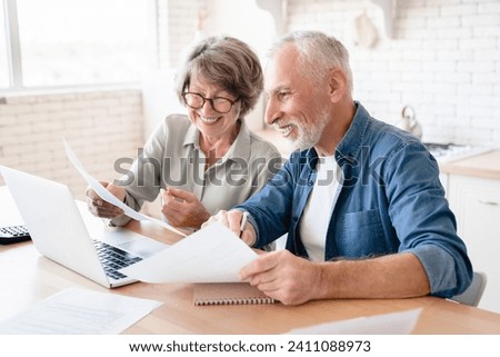 Smiling caucasian old elderly senior couple husband and wife using laptop and doing paperwork together at home kitchen. E-banking, e-commerce