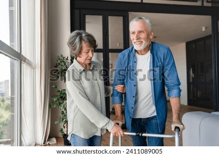 Rehabilitation after traumas injuries. Old elderly senior woman caretaker nurse wife supporting helping disabled handicapped man patient husband to move with walking frame