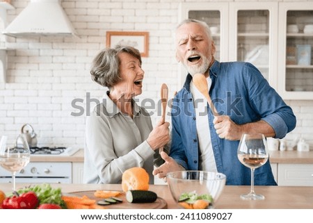 Active seniors concept. Elderly senior old couple grandparents singing dancing at home kitchen while cooking dinner lunch, drinking wine and celebrating special event together Royalty-Free Stock Photo #2411089027