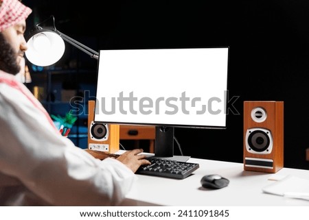 Image portrays Muslim guy seated at his workstation at home, his computer monitor showing an isolated chromakey template. Youthful Arab man working with a desktop pc displaying copyspace white screen. Royalty-Free Stock Photo #2411091845