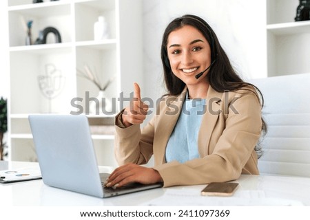 Customer support service. Confident positive woman, call center operator with headset, working on support hotline in a modern office with a laptop, looks at the camera, smiling, shows thumb-up gesture Royalty-Free Stock Photo #2411097369