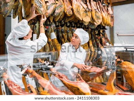 Positive young female and male vendors working at counter in butcher shop, arranging dry-cured spanish jamon for sale Royalty-Free Stock Photo #2411108303