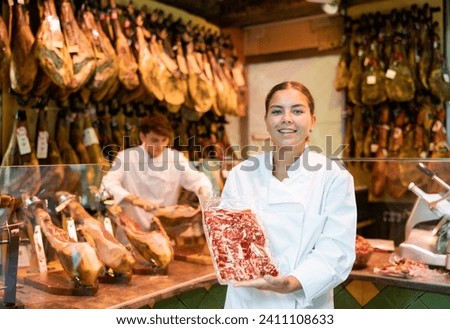 Young female seller offering sliced bacon in butcher shop Royalty-Free Stock Photo #2411108633