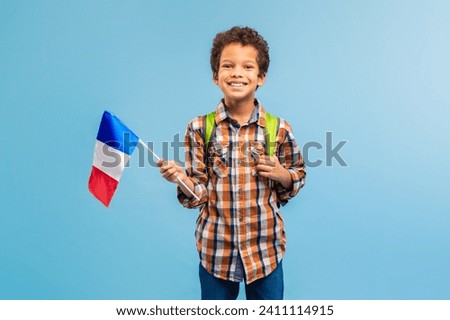 Joyful boy holding French flag, dressed in plaid shirt with backpack, embodying youthful spirit and cultural pride on a blue backdrop Royalty-Free Stock Photo #2411114915