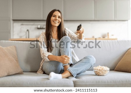 Delighted young female with bowl of popcorn at home comfortably watching her favorite tv shows, smiling woman holding remote in hand, relaxing on plush couch in living room, copy space Royalty-Free Stock Photo #2411115113