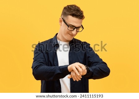 Time management concept. Positive young guy in eyeglasses checking time, looking at smart watch on his wrist and smiling, isolated on yellow studio background Royalty-Free Stock Photo #2411115193
