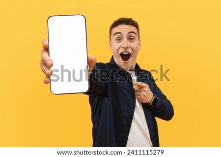 Amazed cool young guy showing big phone with white blank screen and grimacing, recommending nice online offer, deal, mobile app, mockup, copy space, yellow background Royalty-Free Stock Photo #2411115279