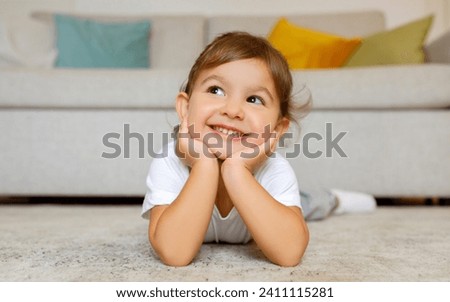 Cheerful cute little girl daydreaming on the floor at home, adorable toddler female child lying on carpet in living room, resting head on hands, smiling and looking away, copy space Royalty-Free Stock Photo #2411115281