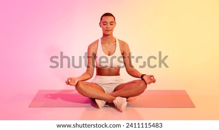 Serene latin young woman with a short haircut in sportswear meditating in lotus position on a yoga mat, eyes closed, against a peaceful pink and yellow gradient background Royalty-Free Stock Photo #2411115483