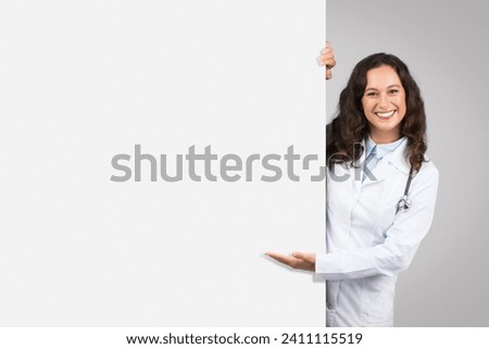 Cheerful female doctor with stethoscope peeking out from behind blank white board, presenting space for text or graphics, grey background Royalty-Free Stock Photo #2411115519