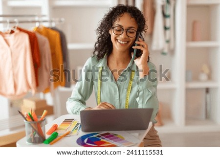 Engaged fashion designer in glasses conversing on phone while working on her laptop, surrounded by vibrant color samples in her studio Royalty-Free Stock Photo #2411115531