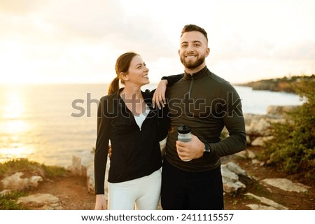 Joyful man and woman in activewear, sharing light moment holding water bottle and embracing against sunset backdrop at seaside Royalty-Free Stock Photo #2411115557