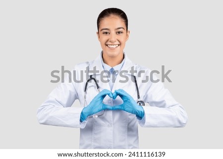 Smiling millennial caucasian doctor, nurse cardiologist in white coat, protective gloves, make heart gesture on chest with hands, isolated on gray background. Health care, love sign, cardio