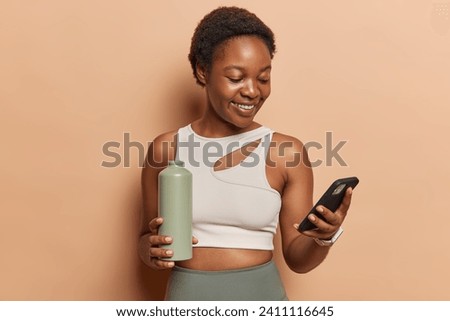 Smiling dark skinned sporty slim woman using smartphone while relaxing after training messaging on cellular hold sport bottle of water dressed in sportswear isolated over brown studio background