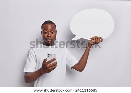 Photo of surprised dark skinned man concentrated in smartphone finds out shocking news holds blank communication bubble for your advertising content isolated over white background. Monochrome shot