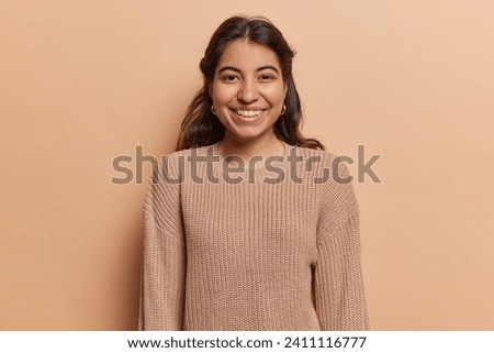 Portrait of pleasant looking Iranian girl with toothy smile has good mood dressed in casual knitted jumper looks directly at camera isolated over brown background. People and positive emotions concept Royalty-Free Stock Photo #2411116777