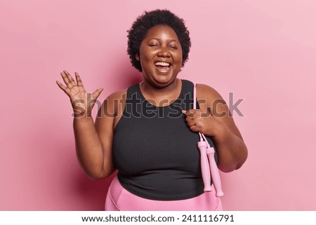 Photo of body positive dark skinned woman enjoys physical activity poses with skipping rope on shoulder happy to loose weight dressed in activewear isolated over pink background. Self acceptance