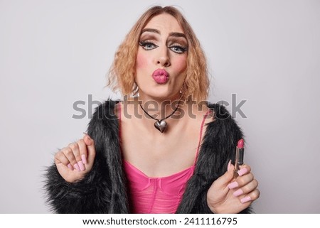 Horizontal shot of gay with vivid makeup keeps lips folded holds lipstick dressed in black fur coat sends air kiss to you has long nails isolated over white wall. Transgender man has feminine style