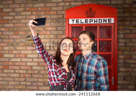 Young Couple Wearing Plaid Shirts Taking Self Portrait with Cell Phone in front of Red Telephone Booth