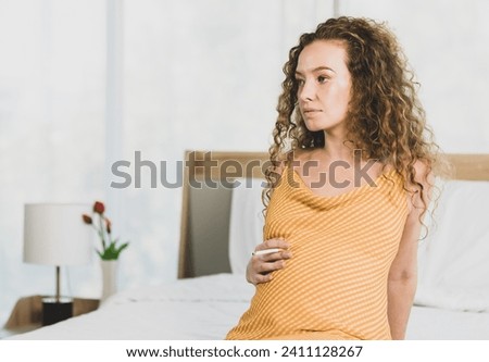 Curly hairstyle young unhappy unhealthy bad behavior Caucasian pregnancy mother in maternity long dress cloth sit on bed in bedroom holding smoking cigarette taking risk and danger to unborn child. Royalty-Free Stock Photo #2411128267