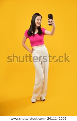 Full body length fun young woman of Asian  in casual clothes doing selfie shot on mobile cell phone post photo on social network isolated on plain yellow background studio portrait