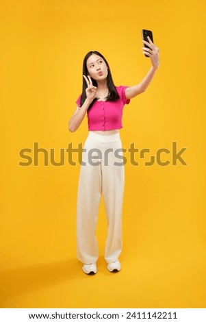 Full body length fun young woman of Asian  in casual clothes doing selfie shot on mobile cell phone post photo on social network isolated on plain yellow background studio portrait