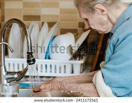 Grandmother washes dishes in the kitchen at home. An old woman does household chores. Washing cups and plates in the kitchen sink. Social roles.
