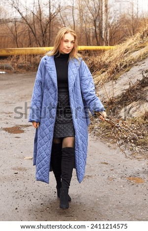 Caucasian young blonde woman with a long blue coat walks along the road with a bouquet of branches of a blooming willow, weather outside the city in spring, symbol of Easter, stylish outfit of a girl
