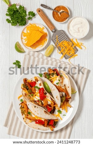 baked tex-mex chicken fajitas with mixed sweet pepper, onion, sour cream, shredded cheese, and white corn tortillas on white plate on wooden table with ingredients, vertical view, flat lay