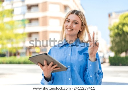 Young pretty blonde woman holding a tablet at outdoors smiling and showing victory sign