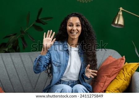 Young beautiful woman at home on sofa looking at web camera, talking with friends remotely, smiling with satisfaction and gesturing with hands, using video call on computer, waving hand greeting. Royalty-Free Stock Photo #2411275849