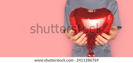 Banner with woman hold in hands red inflatable foil balloon in a heart shape in front of pink background.