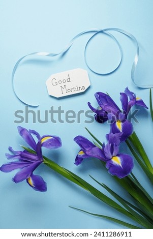 Beautiful purple iris flowers and Good Morning note on light blue background. Flat lay, top view.