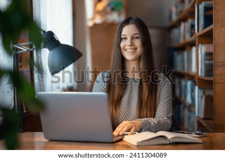Happy university female student in library working at her laptop. Portrait of a smiling girl college student doing assignment in library