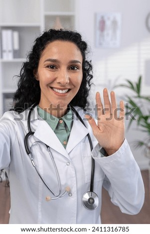 Vertical shot, female doctor smiling joyfully waving at phone camera, talking to patients remotely using smartphone video call app, working inside clinic office. Royalty-Free Stock Photo #2411316785