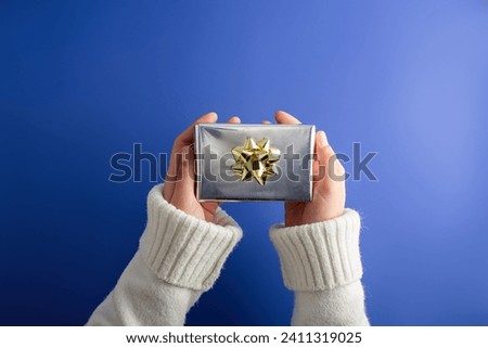 First person top view photo of hands holding shiny silver paper giftbox with vivid golden bow isolated blue background.  Perfect for valentines day marketing


