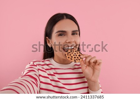 Young woman with chocolate chip cookie taking selfie on pink background