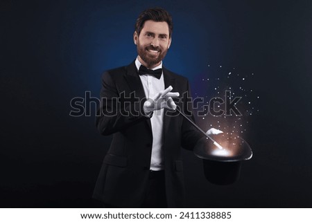 Smiling magician showing trick with top hat and wand on dark blue background