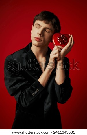 sentimental man in velvet blazer holding heart-shaped present on red background, Valentines day Royalty-Free Stock Photo #2411343531