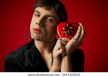sentimental man in velvet blazer holding heart-shaped gift box on red background, Valentines day Royalty-Free Stock Photo #2411343533